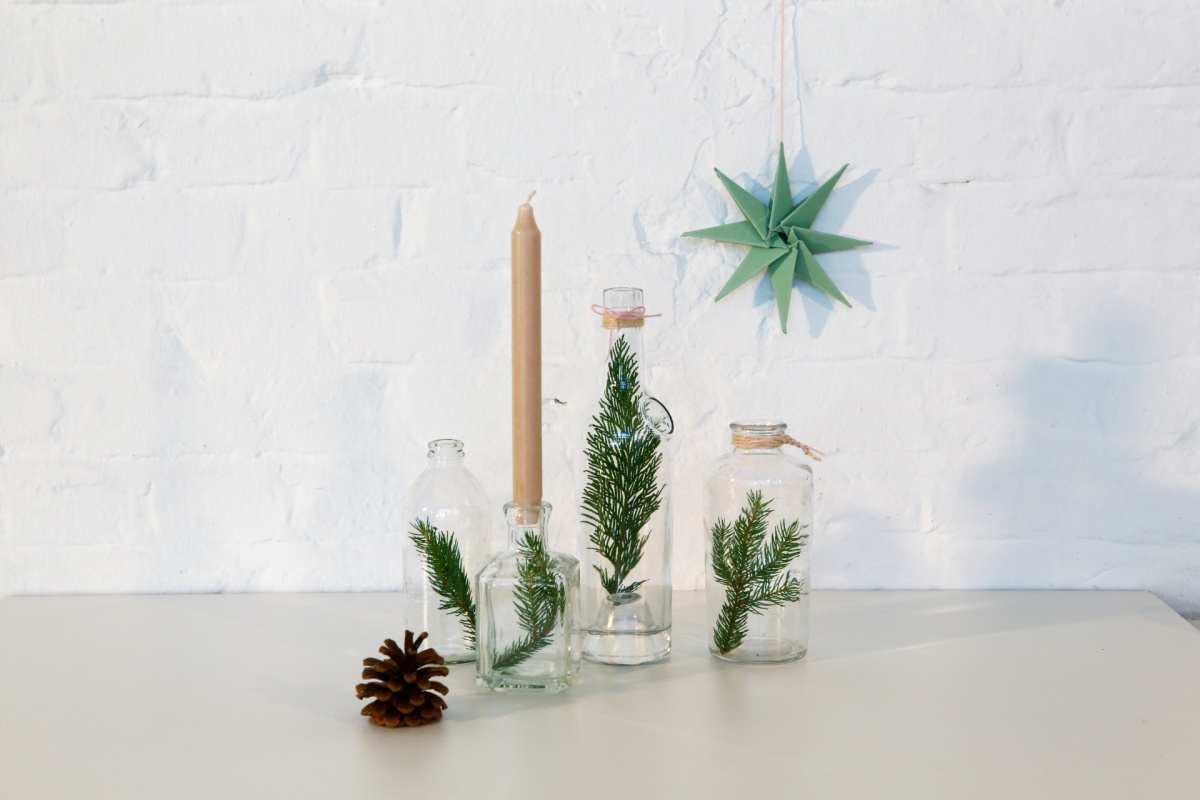 Minimalistische Weihnachtsdekoration mit Tannenzweigen in Glasflaschen, Kerze und gefaltetem Papierstern.