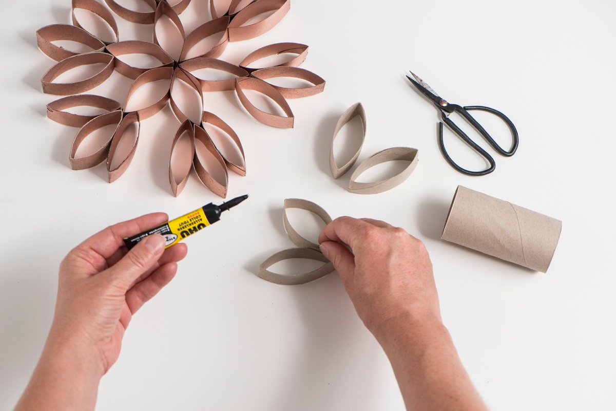 Hände basteln Schneeflocken-Deko aus Toilettenpapierrollen und Kleber, Schere im Hintergrund.