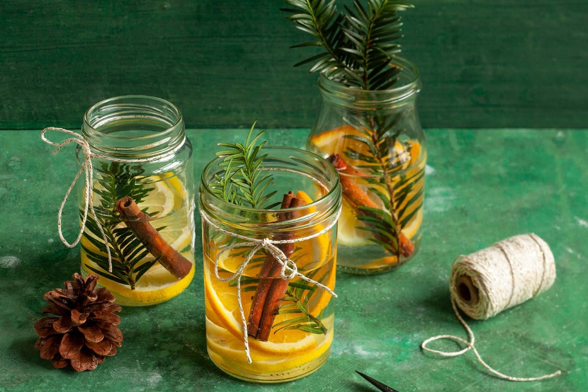 Einmachgläser mit Orangen, Zimtstangen und Tannenzweigen als festliche Tischdeko, umgeben von Garn und Tannenzapfen.