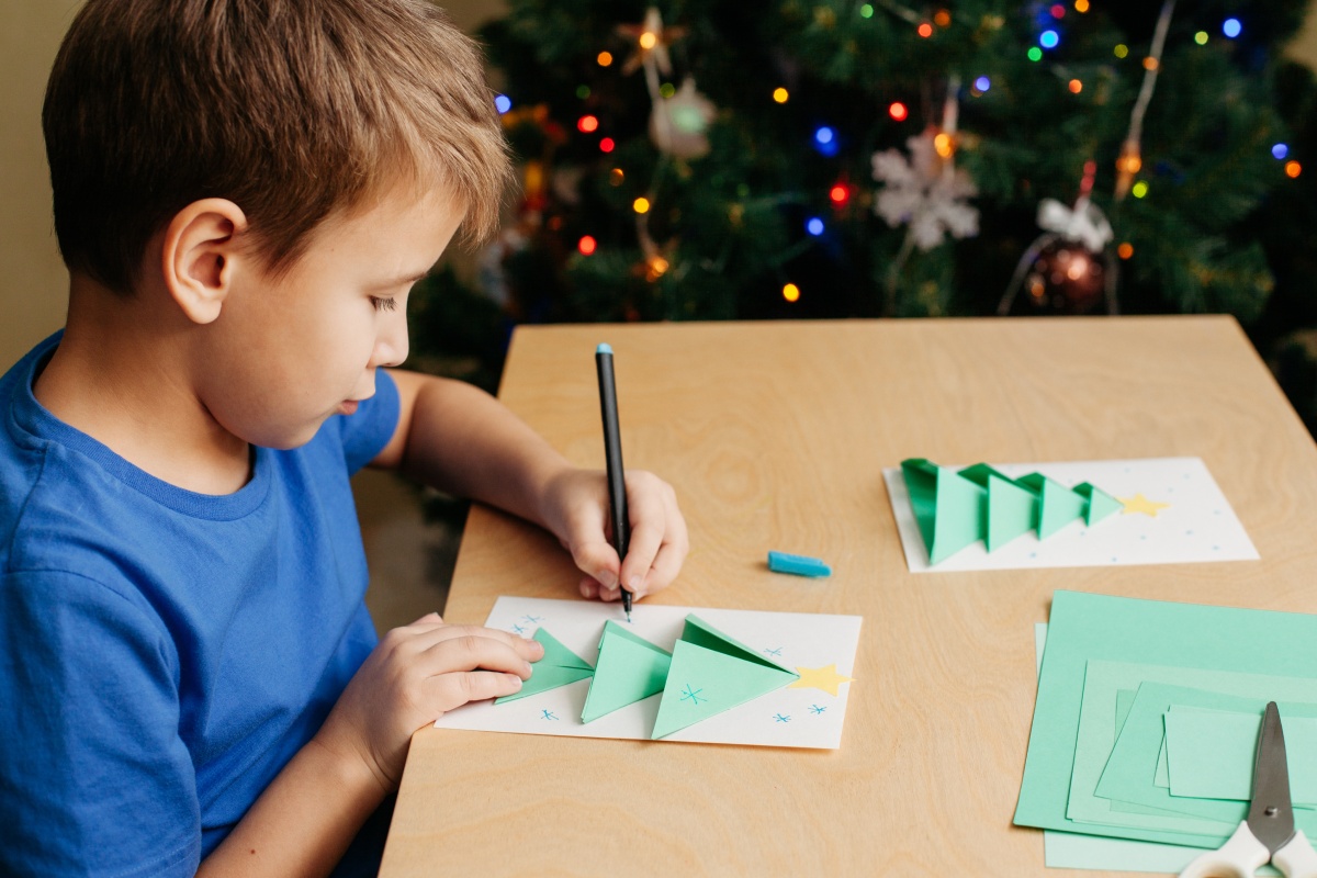 Junge bastelt Weihnachtskarte mit grünem Papier am Tisch vor geschmücktem Weihnachtsbaum.