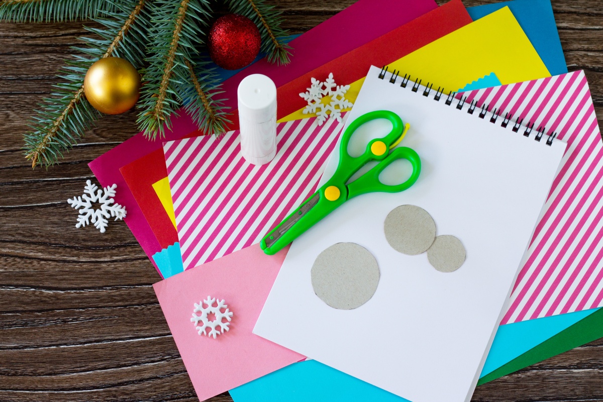 Bastelmaterialien mit Schere, Klebestift, farbigem Papier und Schneeflocken-Dekoration auf Holztisch.