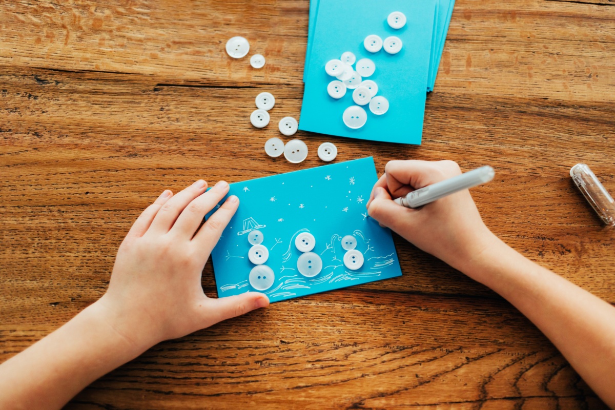 Kind bastelt Schneemannkarte aus blauen Knöpfen und zeichnet Details mit weißem Stift auf blauem Papier.