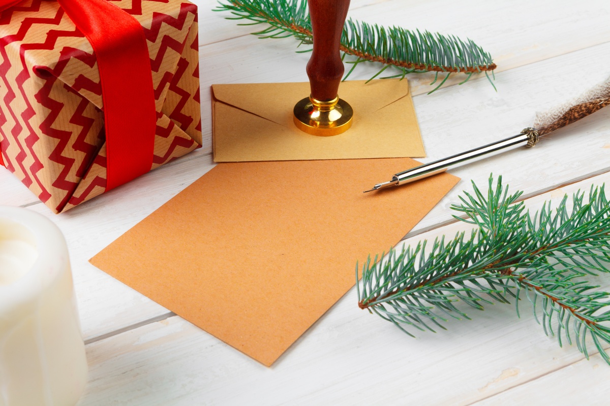 Weihnachtlich verpacktes Geschenk, Brief mit Siegelstempel, Tannenzweige und Federkiel auf hellem Holztisch.