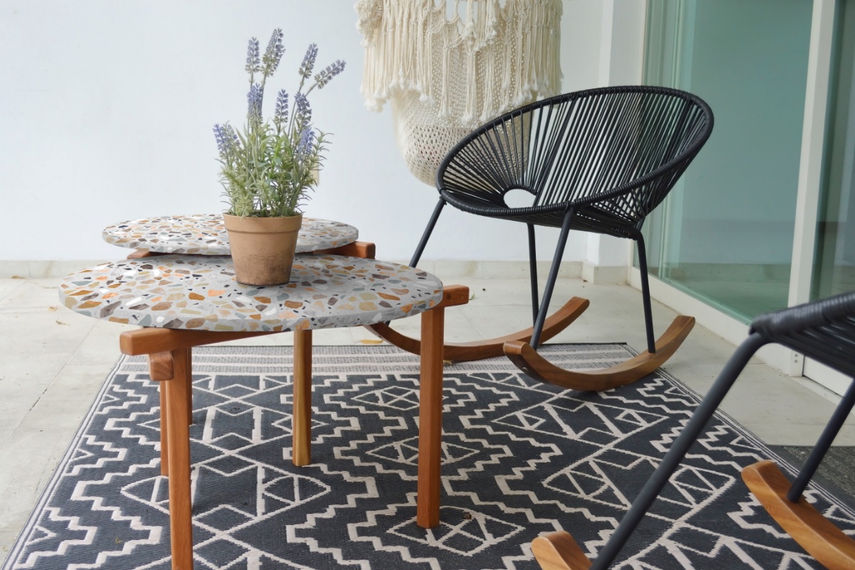 Stilvolle Terrasse mit schwarzem Schaukelstuhl, Terrazzo-Tischen, Lavendelpflanze und Makramee-Deko vor weißer Wand.