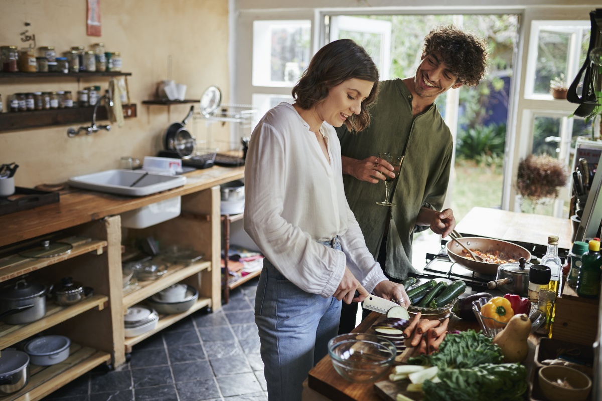 Paar bereitet in einer gemütlichen, rustikalen Küche frisches Gemüse zu, während sie sich unterhalten und ein Glas Wein genießen.