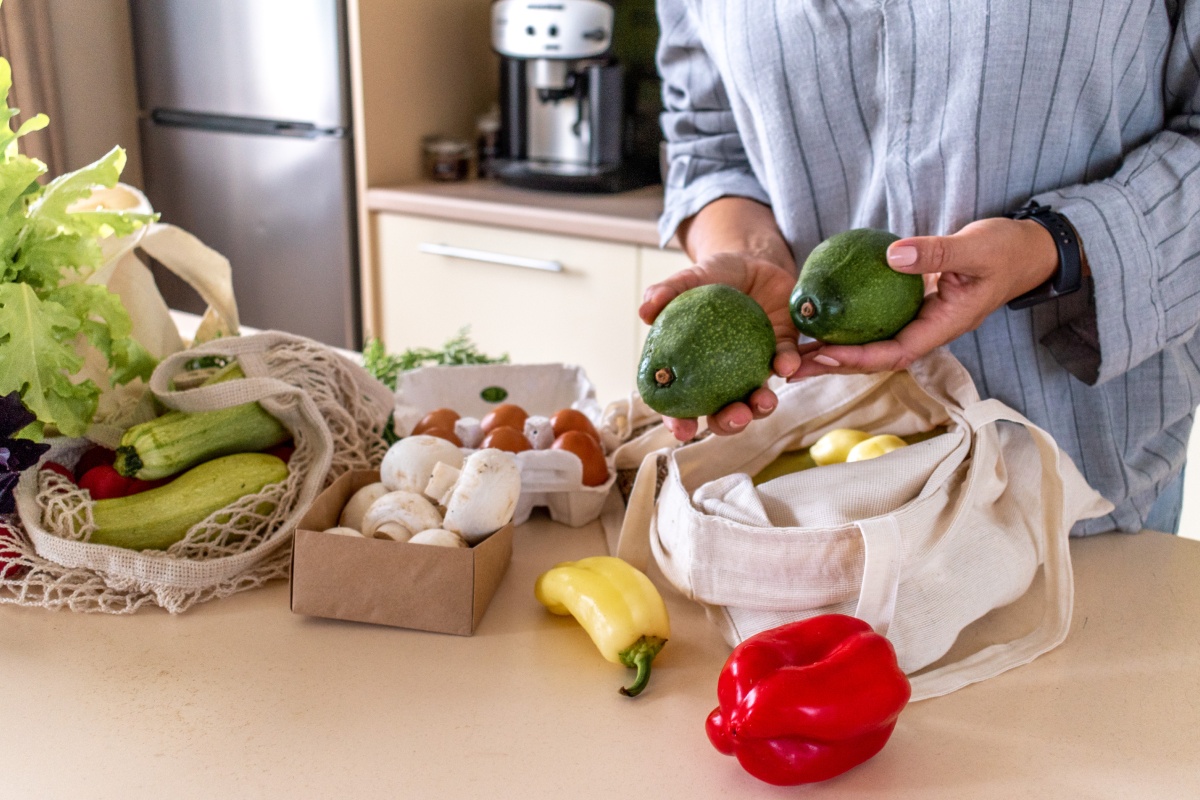 Person hält zwei Avocados über einem Tisch mit frischem Gemüse und Eiern in einer Küche.