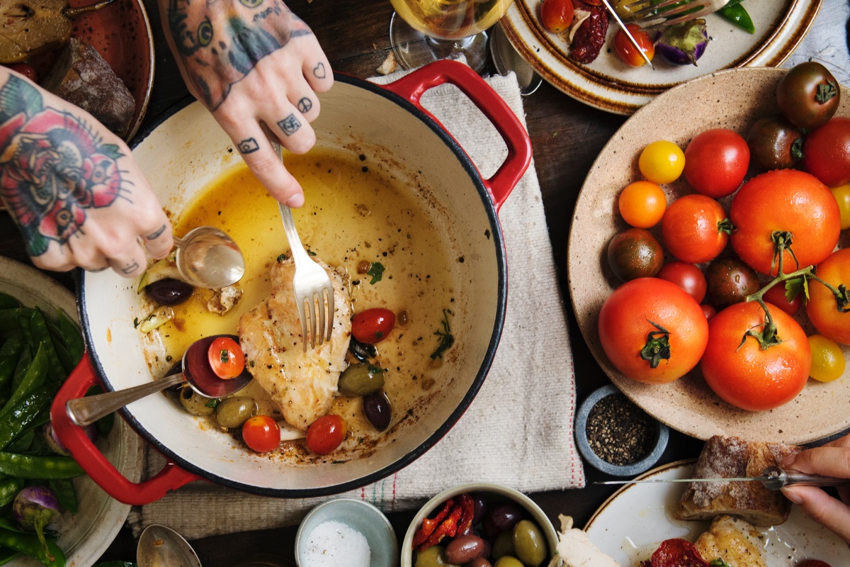 Reich gedeckter Tisch von oben, eine Frau mit tätowierten Händen holt mit einer Gabel eine gebratene Hähnchenbrust mit Tomaten aus einem Brattopf, rechts vom Topf steht eine Schale mit vielen verschiedenfarbigen Tomaten, links vom Topf steht eine Schale mit grünen Bohnen.