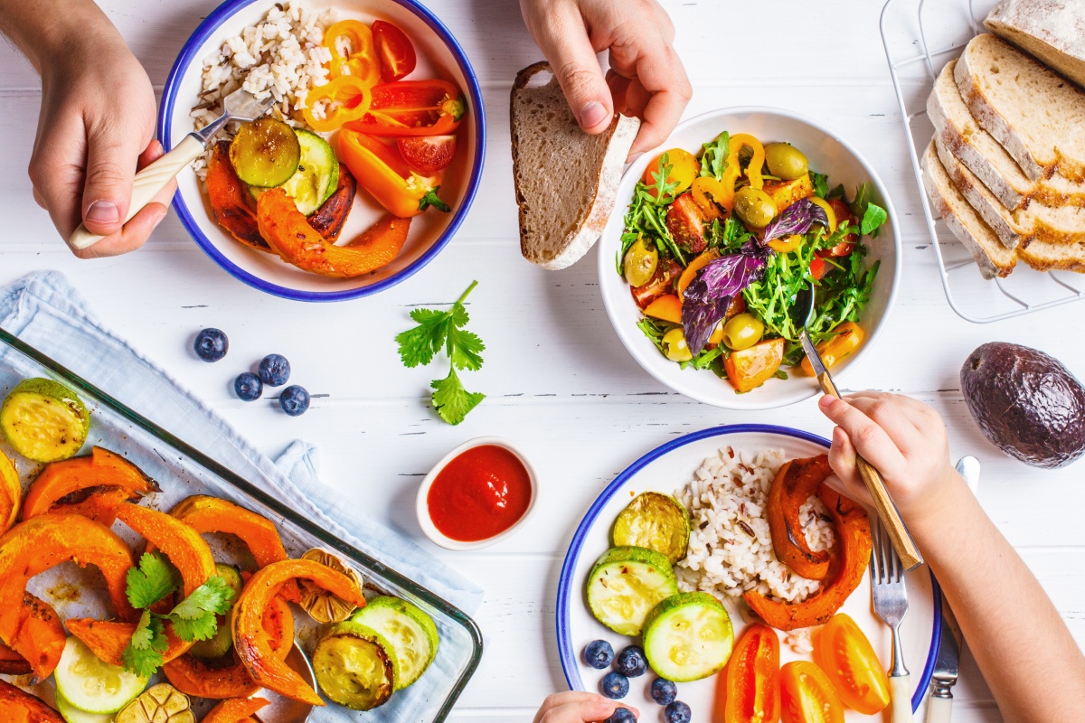 Ein reich gedeckter Tisch mit Tellern und Schüsseln voller Gemüse. Kinder- und Erwachsenenhände bedienen sich.