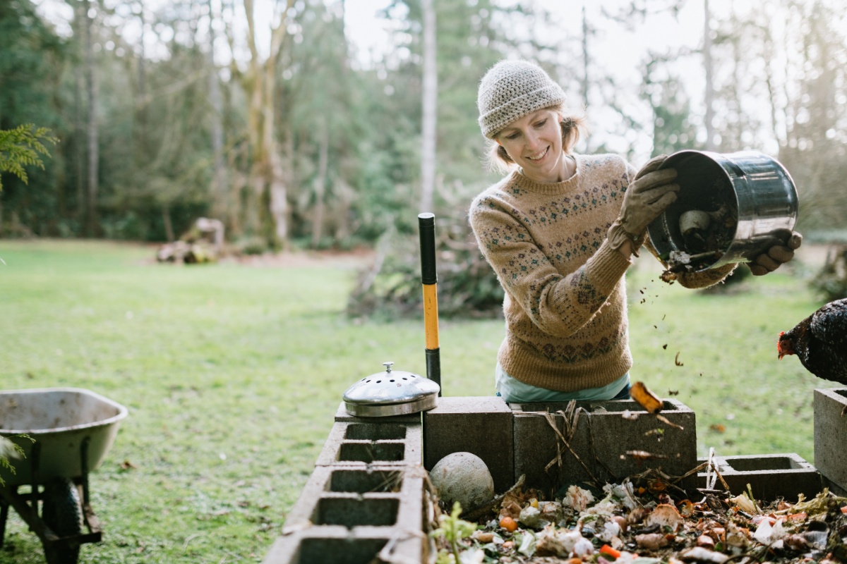 Frau in Mütze, Pullover und Gartenhandschuhen leert Abfalleimer in einen Komposter. Rechts neben ihr ein Huhn, links eine Schubkarre, im Hintergrund ein großer Garten mit hoch gewachsenen Bäumen.