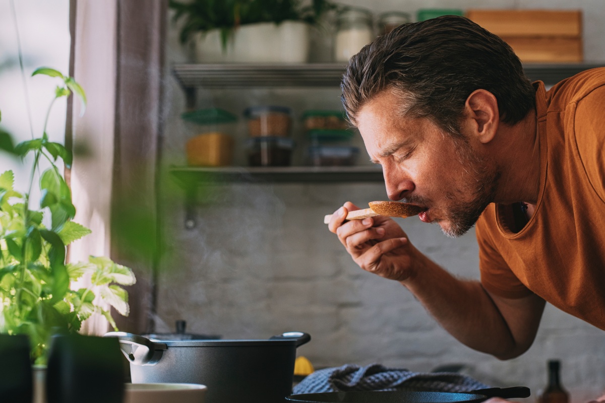 Mann steht in Küche, beugt sich über den Herd mit Töpfen und kostet Tomatensoße vom Holzlöffel