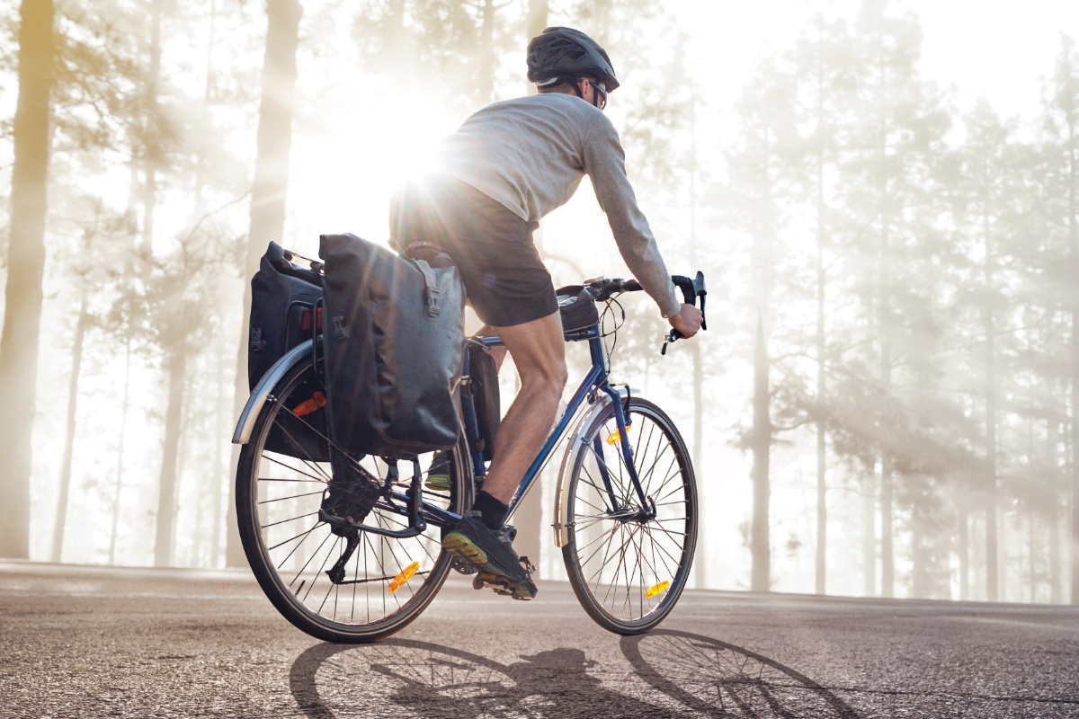 Radfahrer mit zwei Fahrradtaschen am Gepäckträger, Sportschuhe, Radlerhose, Pullover und Helm tragend, fährt eine Bergstraße entlang, rechts von ihm steiniger Hang mit Sträuchern, links Bäume mit einfallenden Sonnenstrahlen.