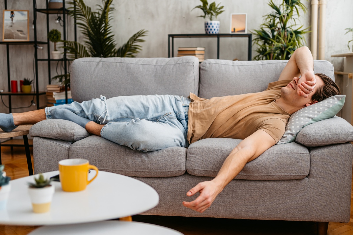 Mann liegt erschöpft auf einer grauen Couch und hält sich die Stirn, typisches Zeichen von Müdigkeit nach dem Essen.