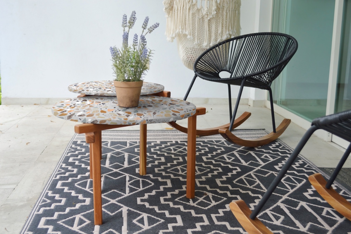 Zwei unterschiedlich große versetzt übereinander angeordnete runde Tischplatten in Terrazzo-Optik mit Tischbeinen aus Holz, darauf eine eingetopfte Lavendelpflanze. Darum stehen zwei runde Rattan-Schaukelstühle und ein Makramee-Hängesessel, auf dem Boden ein gemusterter Teppich.