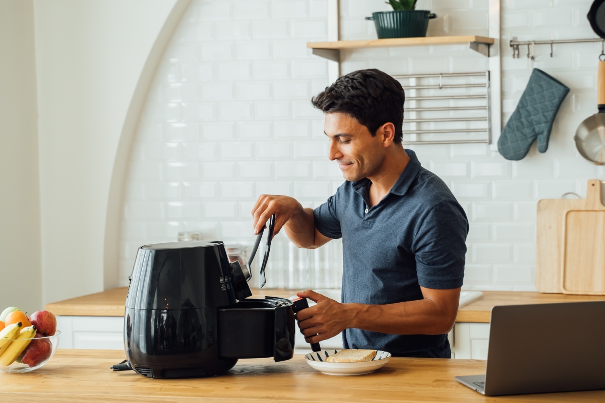 Ein Mann steht mit einer Küchenzange in der Hand vor einer geöffneten Heißluftfritteuse und schaut hinein.