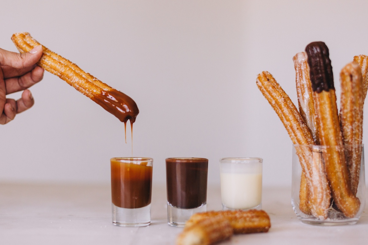 Hand taucht einen Churro in flüssige Schokolade, daneben Gläser mit Karamell und Sahne, weitere Churros im Hintergrund.