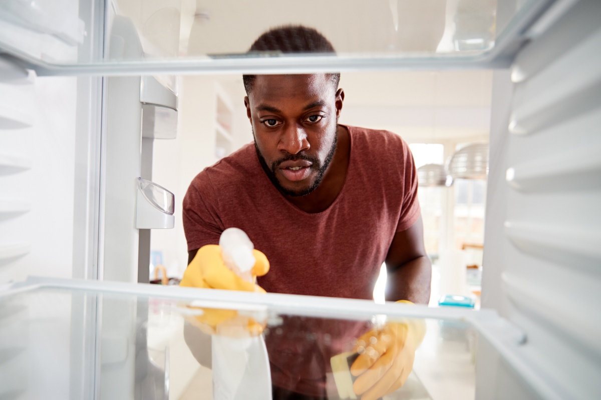 Eine Mann sprüht Reinigungsmittel in den ausgeräumten Kühlschrank.