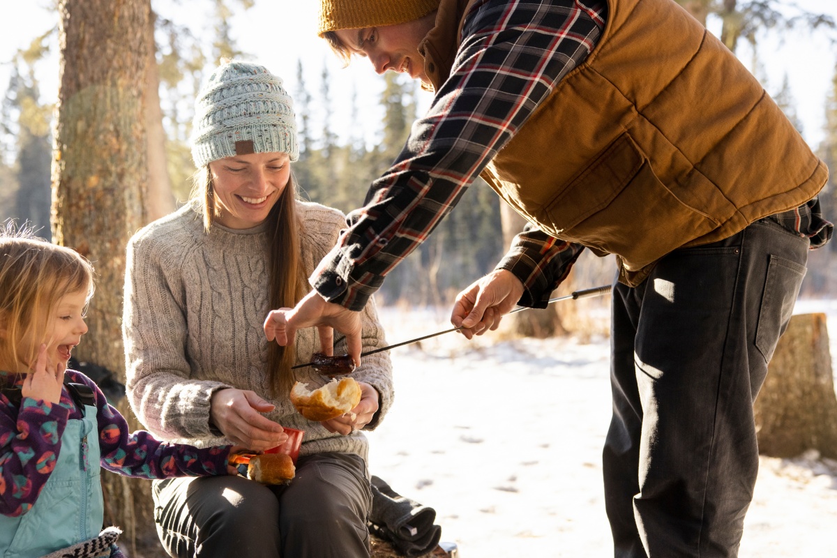 Der Vater legt eine gegrillte Wurst am Spieß auf ein aufgebrochenes Brötchen. Mutter und Kind schauen dabei zu. Im Hintergrund sind Bäume und Schnee zu sehen
