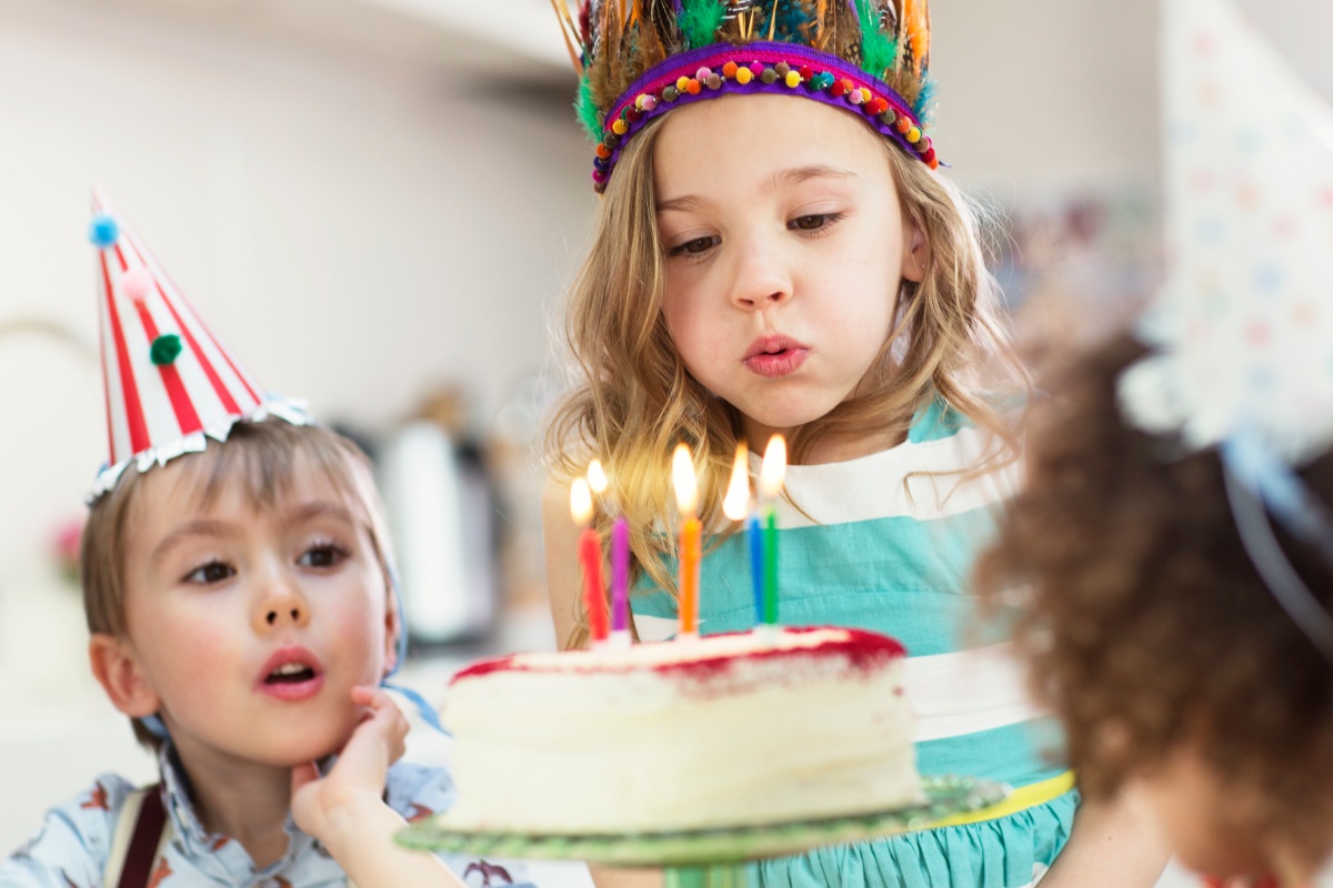 Mädchen mit buntem Feder-Haarreif pustet Kerzen auf einem Kuchen aus. Daneben sitzt ein Junge mit Partyhut und schaut gespannt zu.