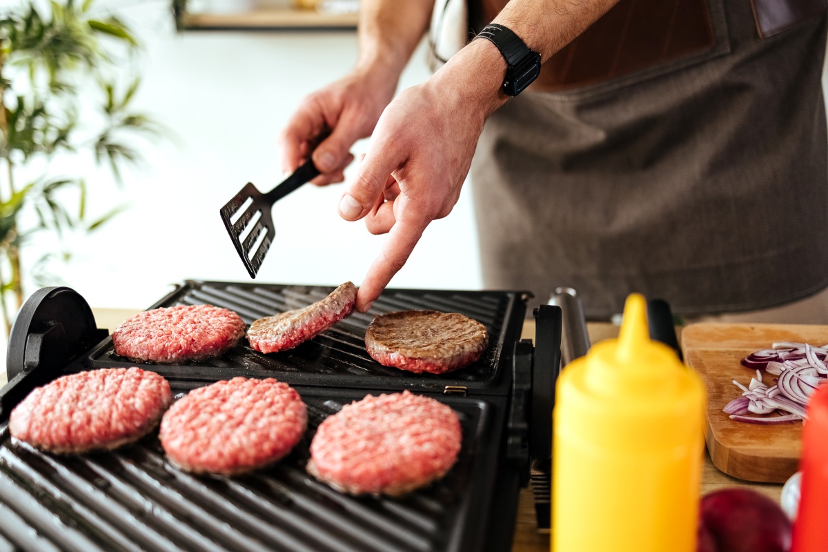 Mann grillt Burger-Patties auf einem Kontaktgrill, gelbe Senfflasche und geschnittene Zwiebeln im Hintergrund.