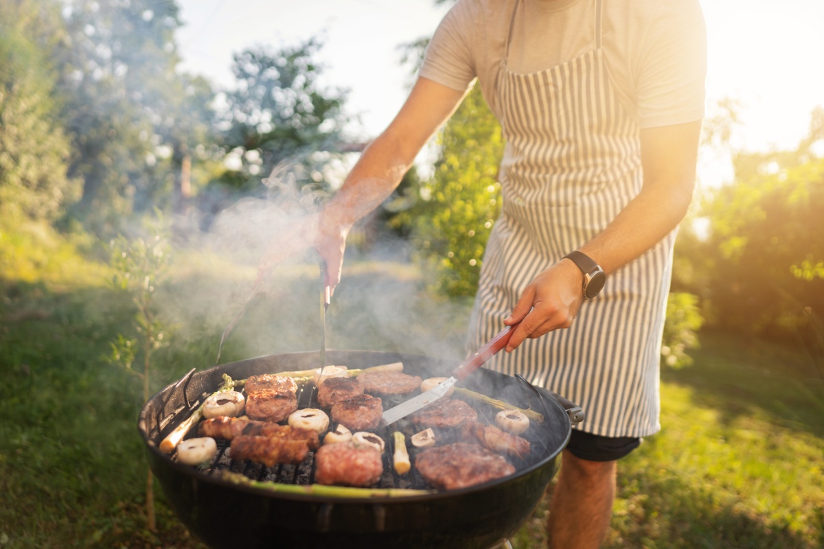 Mann im gestreiften Schürze grillt Fleisch und Gemüse auf einem runden Grill im Garten bei sonnigem Wetter.