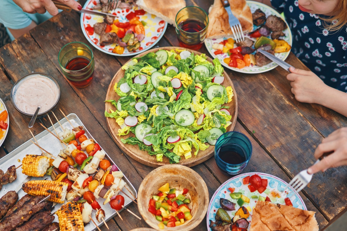 Familienessen im Freien mit Salat, Grillspießen, Gemüse und Getränken auf Holztisch.