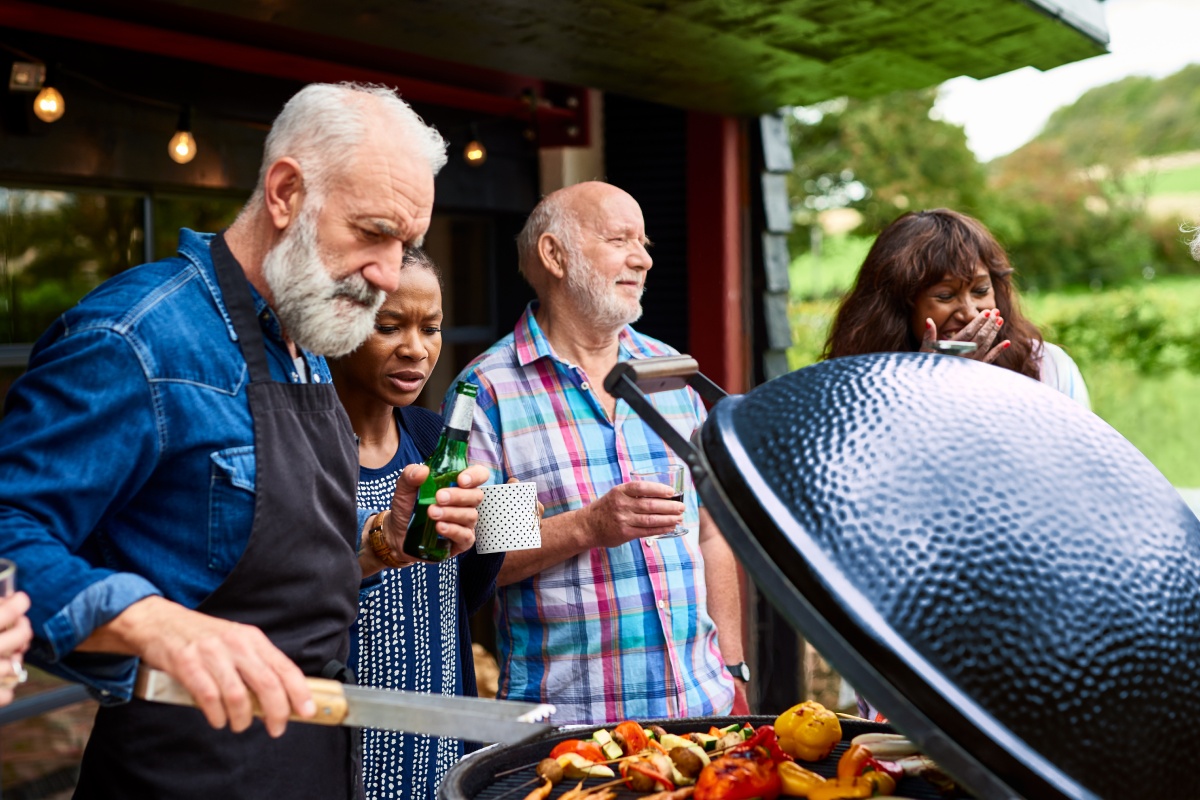 Mehrere Menschen stehen um einen Grill im Garten, trinken was und grillen Gemüse.