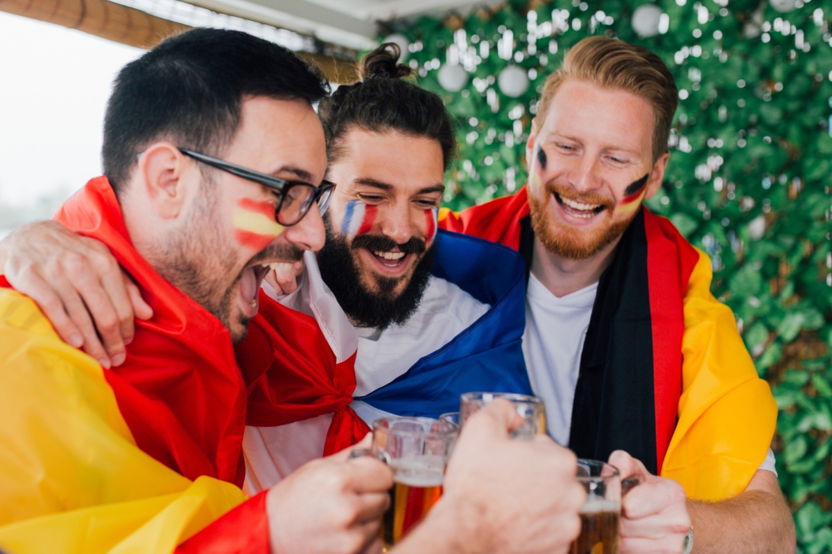 Drei Fußballfans mit bemalten Gesichtern und Flaggen von Spanien, Frankreich und Deutschland stoßen mit Biergläsern an.