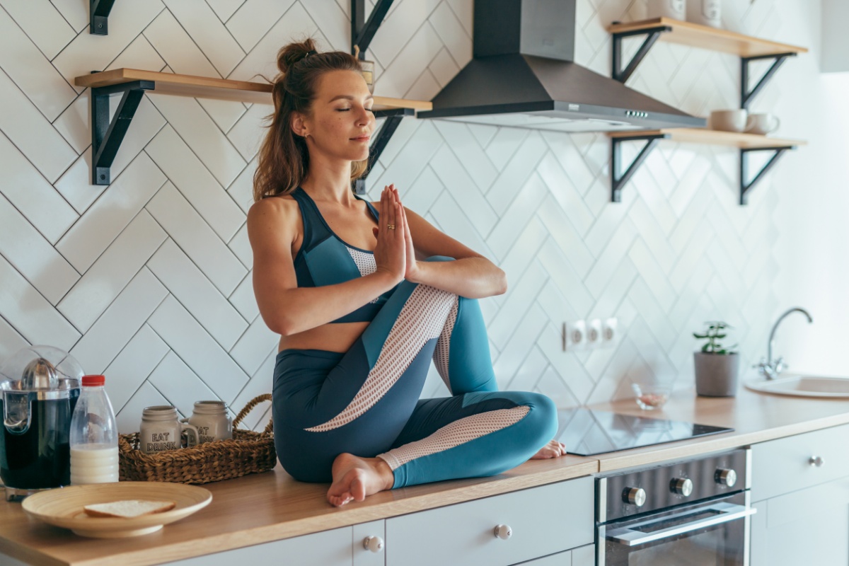 Frau in Leggins und Sport-Bustier sitzt mit geschlossenen Augen in Yogapose mit Händen vor dem Herzen auf einer Küchenzeile.