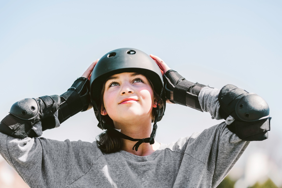 Ein Mädchen mit weißem Sporthelm und schwarzen Ellenbogenschützern schaut zufrieden in den blauen Himmel