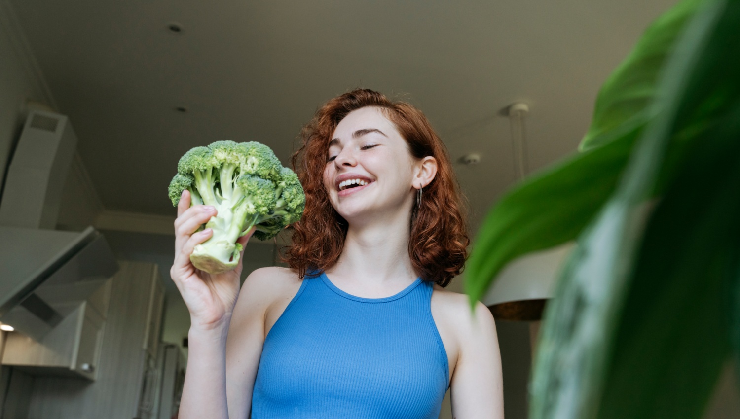 Lächelnde Frau in blauem Top hält einen Brokkoli in der Hand, während sie in ihrer Küche steht.