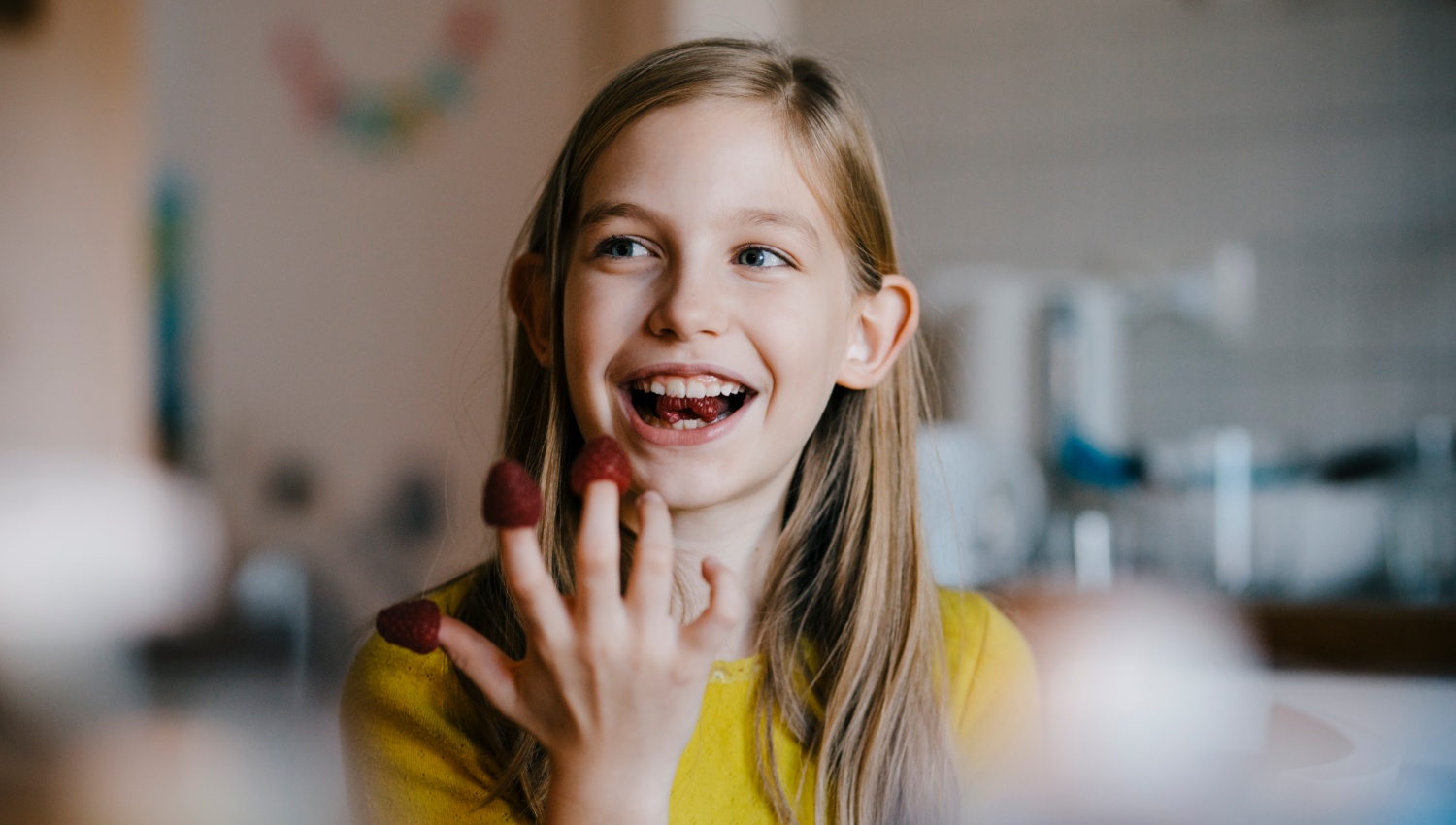 Mädchen isst lachend Himbeeren, die auf ihren Fingern stecken. Freude an guten Snacks in der Kinderernährung.
