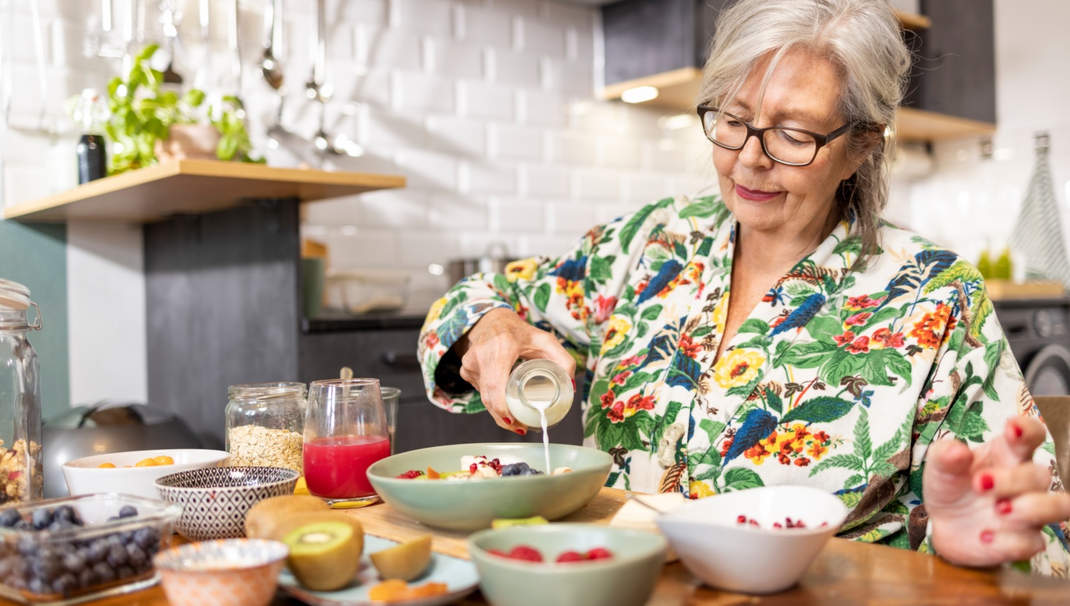 Ältere Frau in einer Küche gießt Milch in eine Schüssel mit Müsli und frischem Obst.