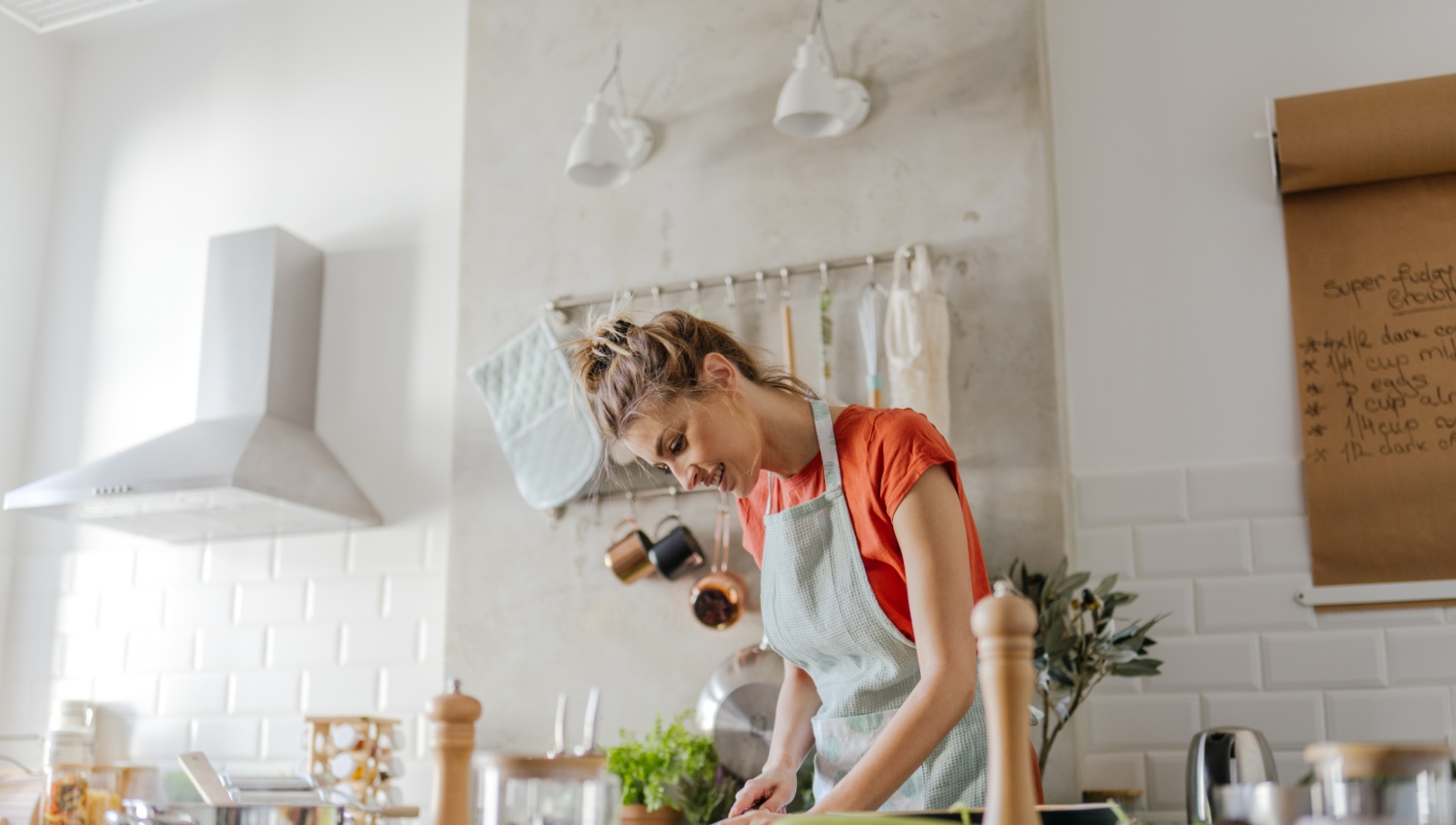 Junge Frau mit Kochschürze  in großer moderner Küche schneidet Gemüse, umgeben von frischen Zutaten.
