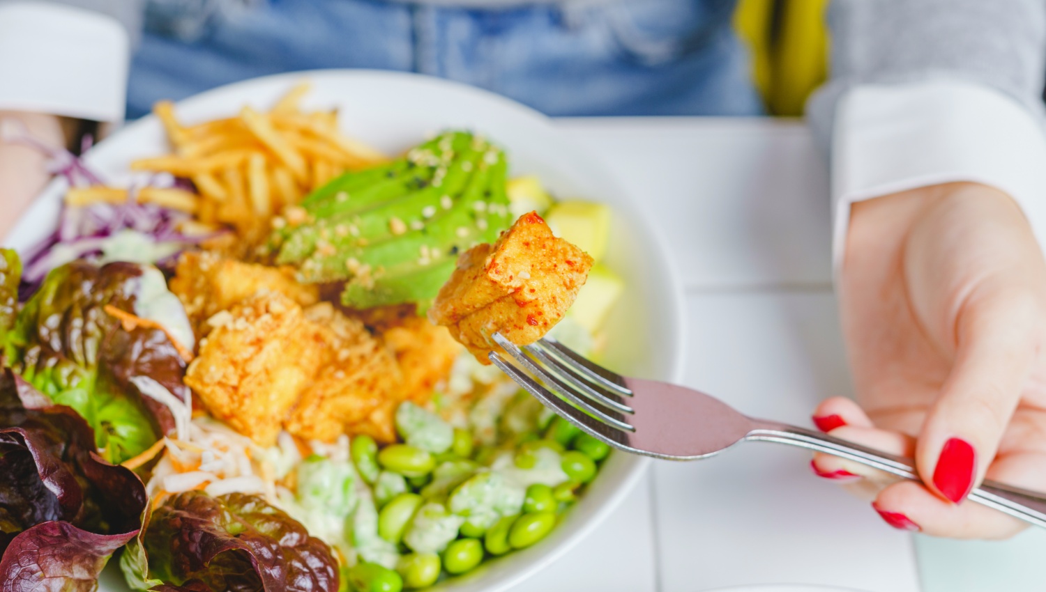 Gabel mit Tofu-Stück über einer farbenfrohen Salatschüssel mit Avocado, Edamame, Pommes und Blattsalat.