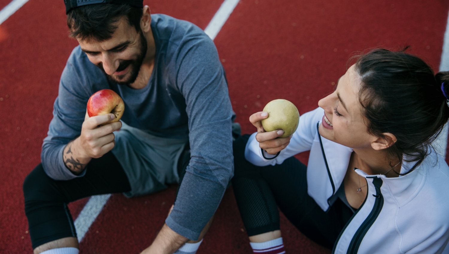 Zwei Personen in Sportkleidung sitzen auf einer Laufbahn und essen lächelnd Äpfel nach einem Training.