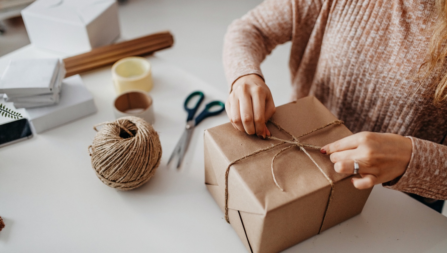 Frau verpackt Geschenk in braunem Papier mit Juteschnur, umgeben von Verpackungsmaterialien.