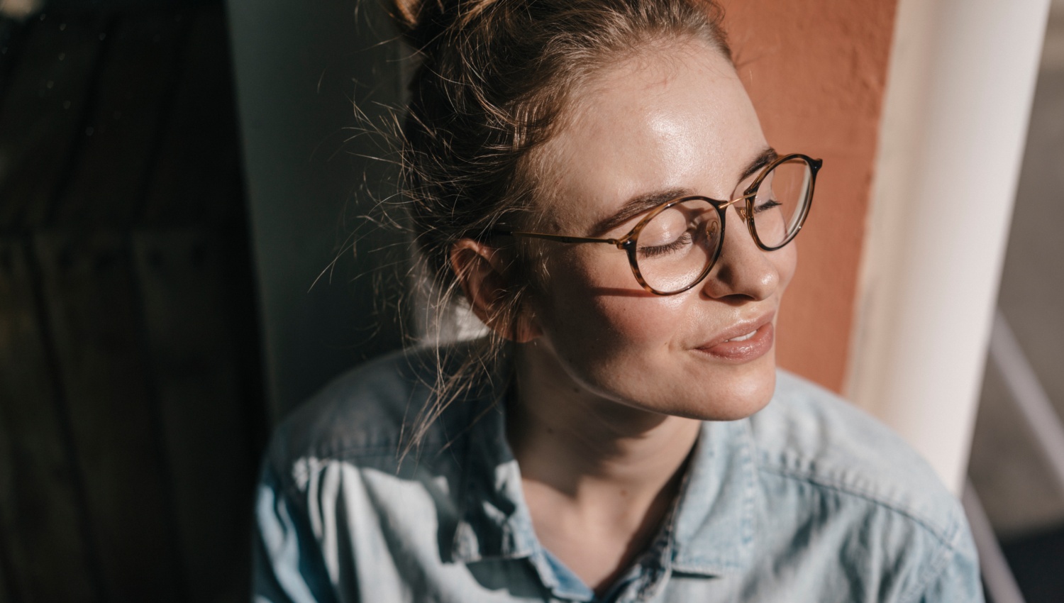 Junge Frau mit Brille schaut mit geschlossenen Augen aus dem Fenster, während die Sonne auf ihr Gesicht scheint.