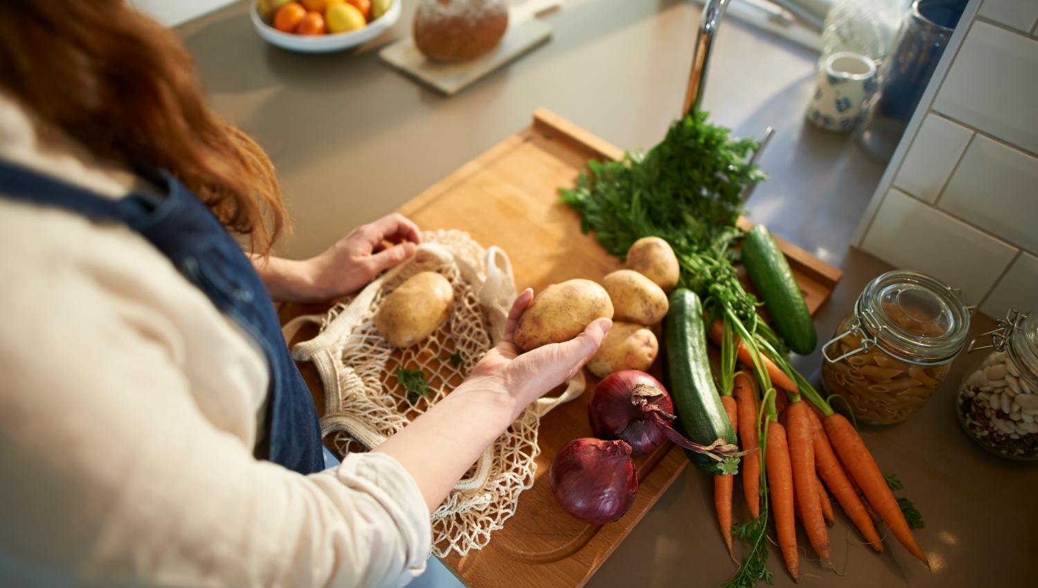 Eine Frau legt Kartoffeln aus einem Netzbeutel auf eine Küchenarbeitsplatte mit frischem Gemüse.