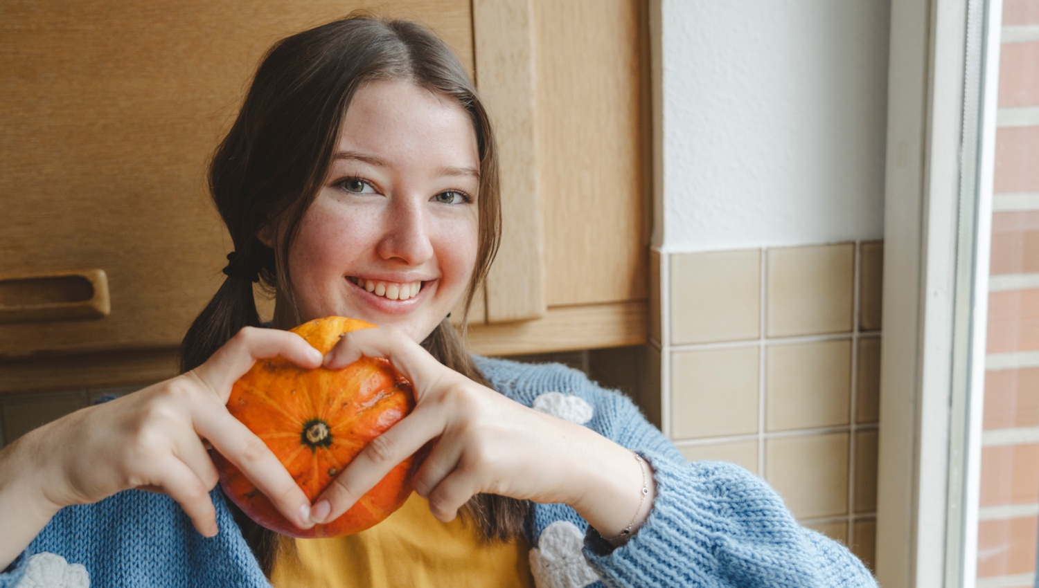 Lächelndes Mädchen hält einen kleinen, orangefarbenen Kürbis und formt ein Herz mit den Händen.