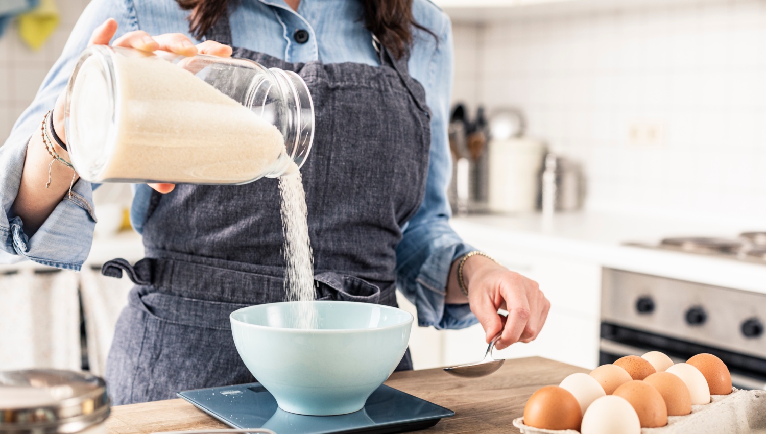 Frau in Schürze gießt Zucker in eine Schüssel, umgeben von Backzutaten wie Eier und Butter.