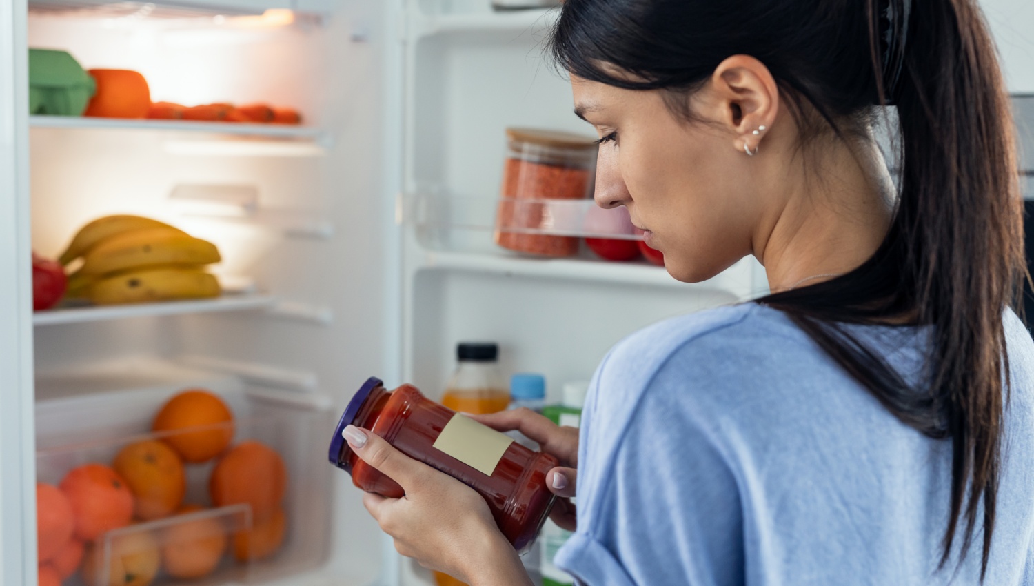 Frau prüft ein Glas Marmelade auf Haltbarkeit vor einem offenen Kühlschrank.