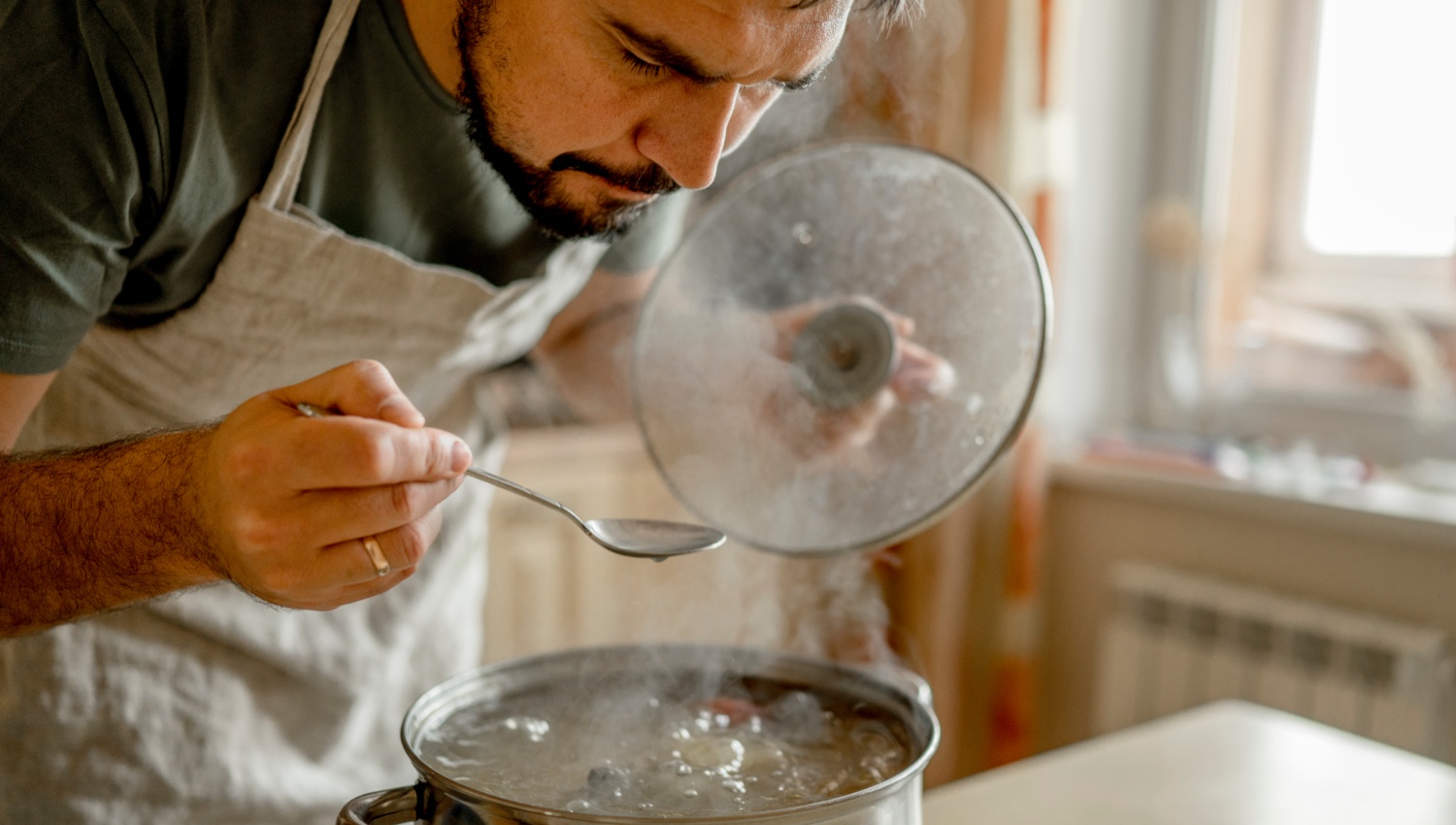 Mann in Schürze probiert Suppe mit einem Löffel aus einem dampfenden Kochtopf.