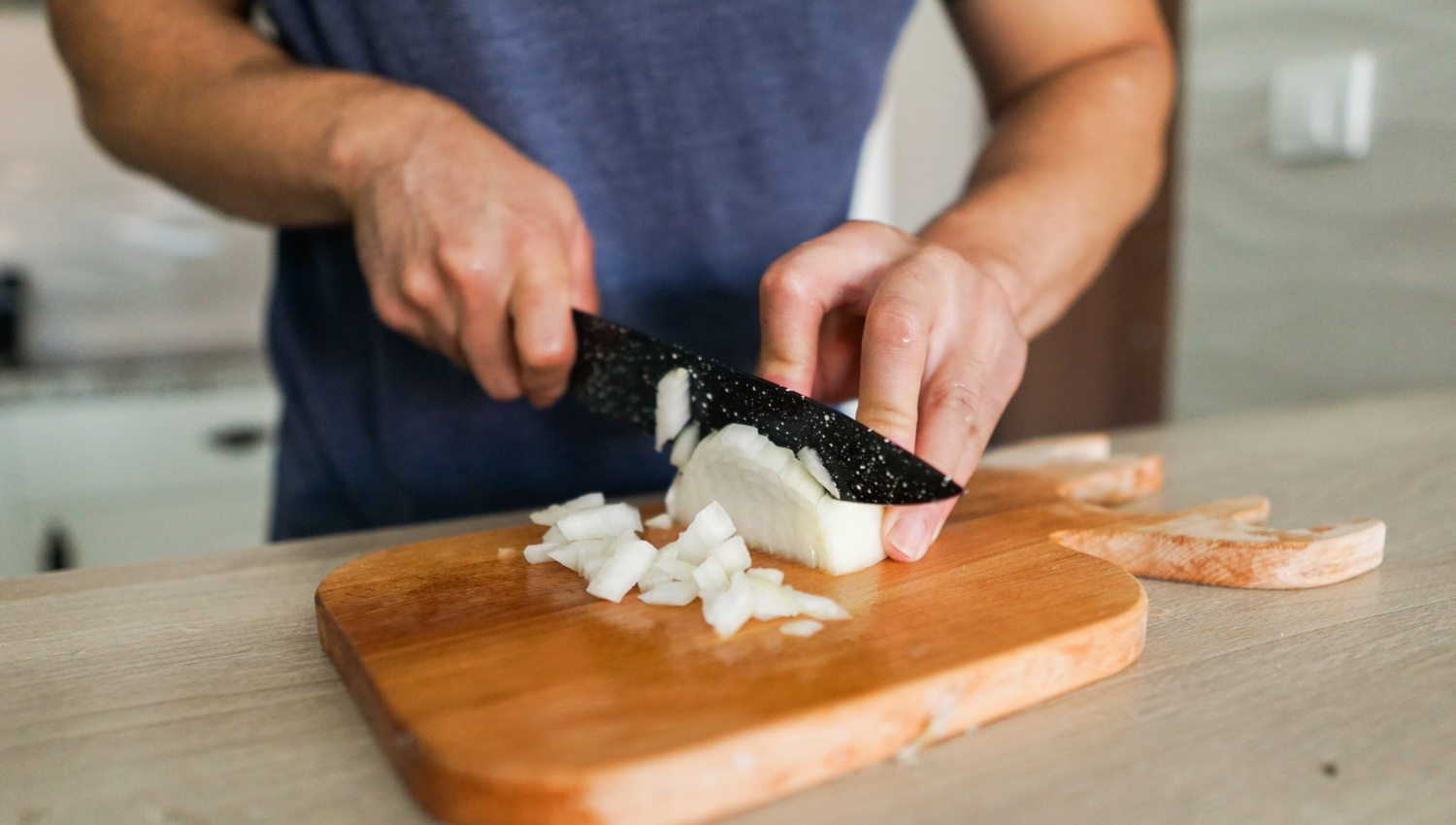 Person schneidet eine weiße Zwiebel auf einem Holzbrett in kleine Würfel.