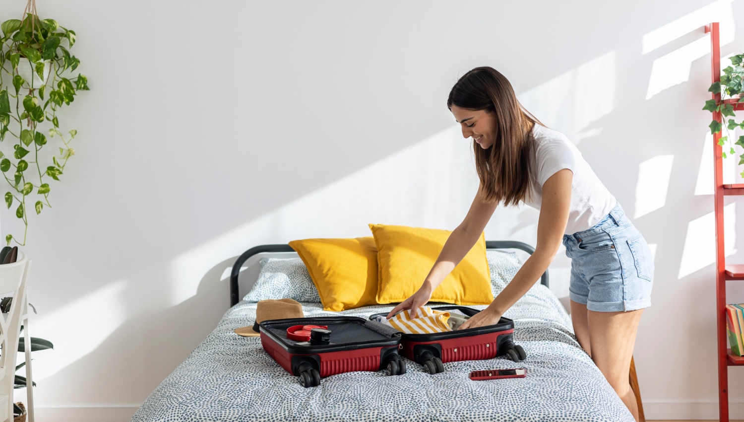 Frau packt einen roten Koffer auf einem Bett mit gelben Kissen in einem hellen, modernen Schlafzimmer.