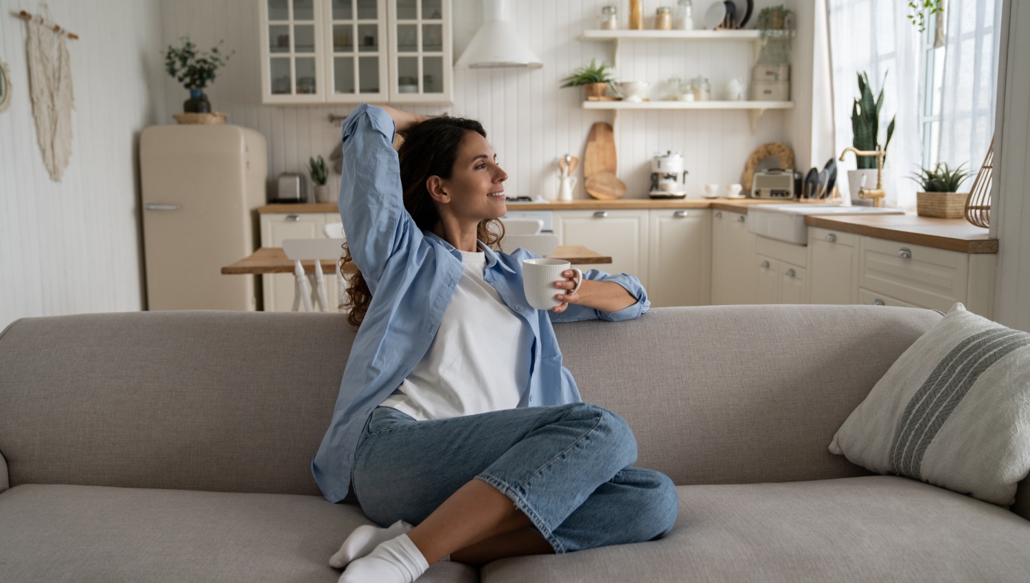Frau sitzt entspannt auf einem Sofa mit einer Tasse Kaffee in einer hellen aufgeräumten Wohnküche.