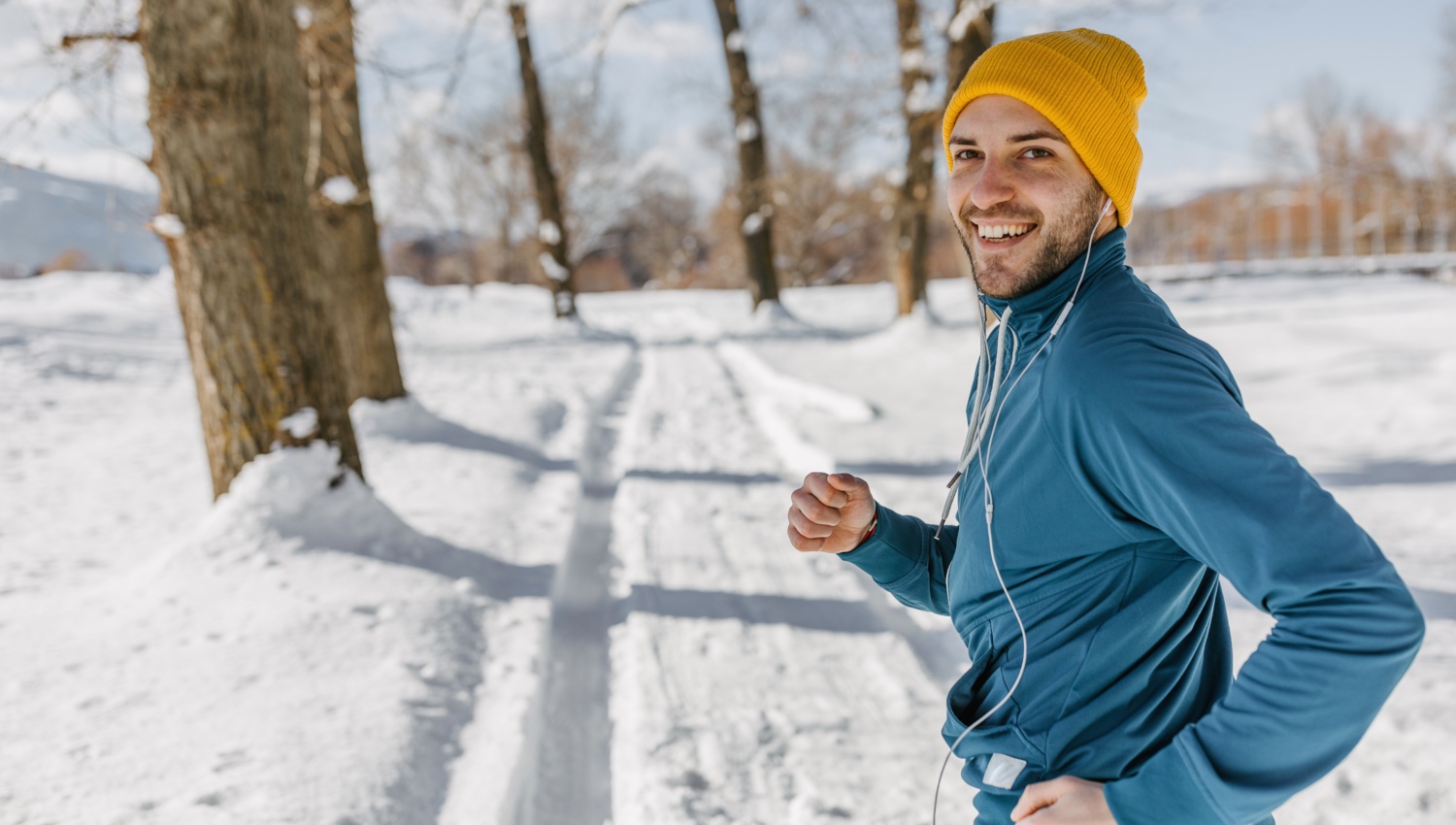 Ein Mann mit Sportkleidung und Mütze joggt lächelnd auf schneebedeckter Laufstrecke im Winter.