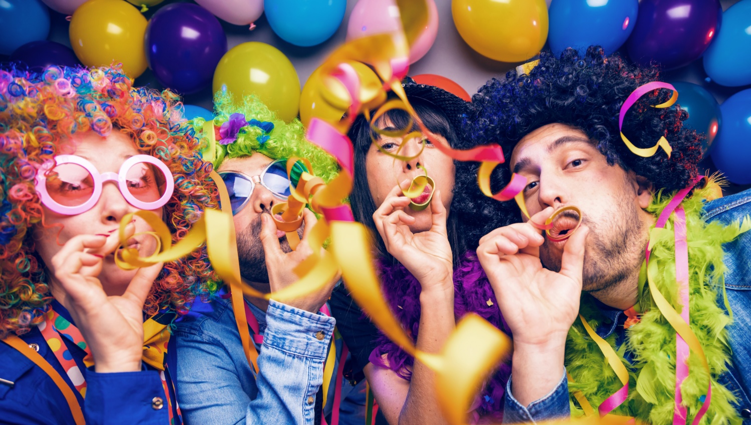4 Personen stehen vor einer Wand mit Luftballons in bunten Faschingskostümen und pusten Luftschlangen in die Luft