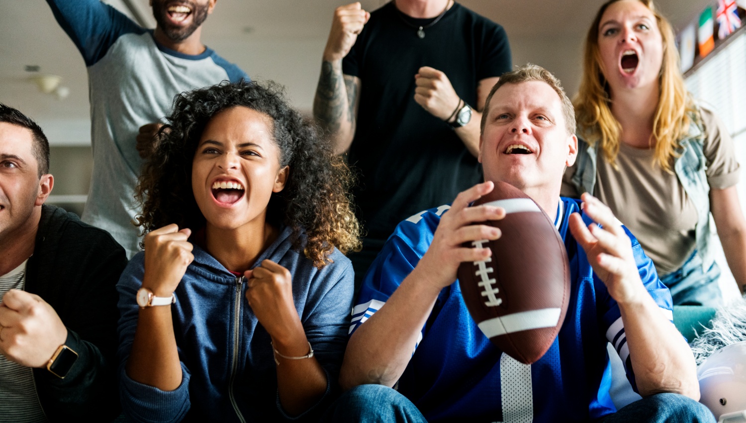Gruppe begeisterter Menschen jubelt beim Ansehen eines American-Football-Spiels in einem Wohnzimmer.