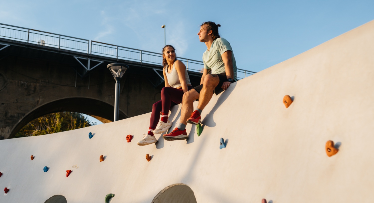 Zwei junge Erwachsene sitzen auf einer Kletterwand im Freien, lächeln und genießen das Sonnenlicht, mit Brücke im Hintergrund.
