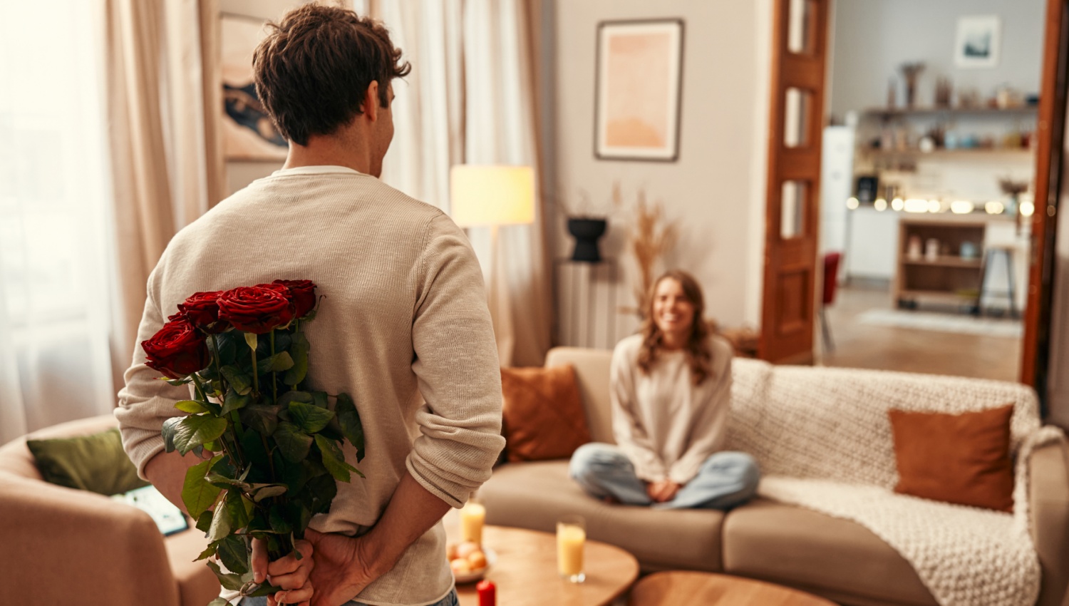 Mann versteckt einen Rosenstrauß hinter dem Rücken, während eine Frau lächelnd auf einem Sofa in einem gemütlichen Wohnzimmer sitzt.